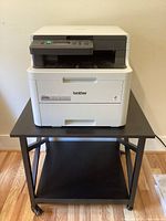 Front view of Brother HL-3290CDW printer on black rolling stand with caster wheels on wooden floor.