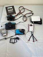 Full view of the lot arranged on a white surface showing Diana F+ cameras, Canon Powershot D20, pink Flip video camera on tripod, Polaroid instant camera, and Polaroid Cube action camera with accessories.