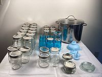 Photo of clear and blue Ball glass canning jars with lids, silicone funnels, and large stainless pot arranged on a table.
