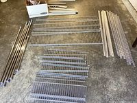 Full view of kit components laid on floor showing metal wire shelves, mounting brackets, and extension rods in silver/chrome finish.