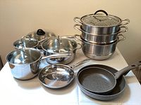 Wide shot of all cookware showing stainless pots, glass lids, two grey Ballarini frying pans and large stainless three-tier steamer.