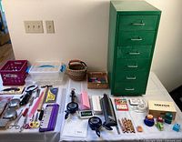 Full lot overview showing green 6-drawer metal storage cabinet and assortment of pens, pencils, scissors, sharpeners, adhesive tape, rulers, and small crafting tools arranged on table