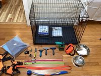 Front view of black metal wire dog crate with plastic base tray, two packages (training pads and crate pads) inside the crate, and multiple dog accessories arranged in front of the crate on the floor. Accessories include two orange harnesses, assorted leashes, grooming tools, stainless steel bowls, small scissors, nail clippers, and a blue ball toy.