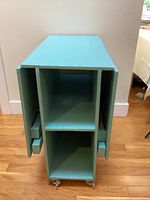 Folded blue painted wooden folding table showing front edge with two vertical storage compartments and shelf inside, on caster wheels.