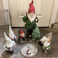 Photo showing all garden gnomes arranged in a semi-circle around round welcome plaque on floor. The tallest gnome has a red hat and green clothes; a green ceramic gnome is also visible.