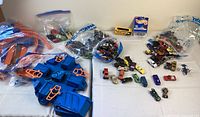 Photo showing blue and orange Hot Wheels track pieces and multiple Hot Wheels cars, some loose and some in bags, laid out on a white surface.