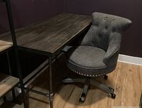 Desk and office chair setup in a room with dark accent walls and wood plank flooring, showing the industrial desk base and the upholstered chair rolled beside it.