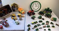 Photo showing a variety of John Deere toy tractors, farm equipment models, and a John Deere themed clock on the wall. Also visible are additional toy trucks and construction vehicles to the left side.