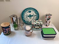 Photo showing all items on table including French press, floral storage tin, ceramic mug and butter dish, insulated tumbler, LunchBots containers, small glass and metal containers, and wall plaque.
