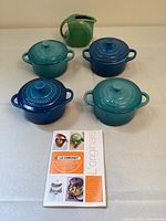 Four Le Creuset mini round cocottes with lids in varying blues and teals, plus a small green ceramic pitcher and a Le Creuset booklet on table.