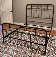 Full view of metal queen size bed frame on patterned rug. Showcases headboard and footboard with pewter/bronze finish and circular centerpiece design.