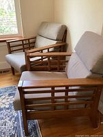 Pair of mid-century style armchairs with wooden frames and beige cushions in a corner of a room with wood floors and windows.