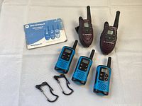 Overview of five Motorla Talkabout walkie talkies, three blue and two purple, laid out with a manual and three clip attachments on a white background.