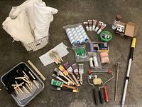 Painting supplies array showing multiple brushes, rollers, drop cloths, and caulking tubes laid on a concrete floor.