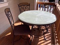 Photo of round patio table with faux green concrete painted wooden top and black iron base, surrounded by three chairs with black metal frames, wooden backs and upholstered seats, placed on a wooden porch.
