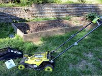 Side view of lime green and black Ryobi 18V battery-powered lawnmower with grass catcher bag, placed on grass near raised garden beds.