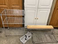 Set of three shelves: large three-tier chrome steel wire shelving unit, long shoe shelf with wooden top, and disassembled steel wire shelf on floor with assembly parts in plastic bin.