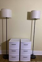 Two white Sterilite 3-drawer plastic storage units with wicker-style front and metal handles positioned between two black metal floor lamps with beige drum shades, against a beige wall.
