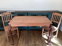 Wooden rectangular table with curved legs and two wooden chairs placed on a wood floor with a green sideboard in background.