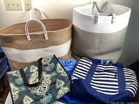 Four large carrying bags displayed together: two woven rope Nautimia-style diaper bags in grey and white, taupe and beige, and two fabric bags (floral and stripe)
