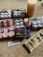 Top view of assorted candles including packages of white flameless tea lights, pink votive candles, colored tealight candles, a large beige pillar candle, and white wax tea lights.