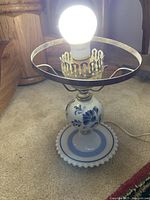 Front side of the vintage porcelain lamp showing the blue floral design on the base and the brass decorative bulb holder with circular metal frame for shade.