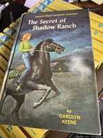 Book cover of 'The Secret of Shadow Ranch' showing a girl on a black horse at night with a cabin and ghostly figure in the background.
