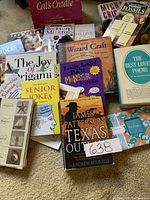 Photo showing assortment of books and a Cats Cradle string game box on carpet, includes titles about jokes, painting, fossils, rocks, poems, origami.