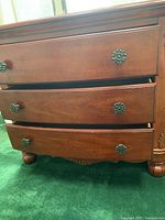 Close-up of one nightstand showing three wooden drawers slightly open with metal floral knobs and carved details on bottom edge and bun foot.