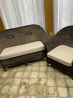 Wicker sofa and chair with cream cushions in a room with tiled floor and yellow walls, front view showing sofa's diamond pattern on backrest and cushion condition.