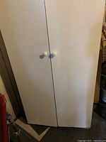 Front view of white two-door storage cabinet with white round knobs, showing some wear marks.