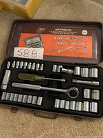 Open metal carrying case showing arranged chrome sockets, ratchet, screwdriver handle, and extensions with an orange label inside the lid showing diagram of set layout.