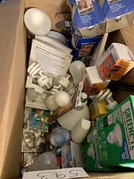 Box containing assorted light bulbs including CFL spiral bulbs, LED bulbs, incandescent bulbs, and various packaging.
