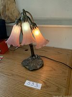 Full view of the lamp on a wood table, showing three pink glass flower shades lit up, with metal base and stems.