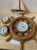 Set of nautical items including a wooden ship wheel clock, two brass instruments, and a model sailboat on a wooden surface.