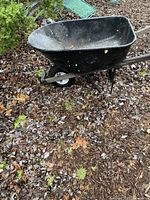 Side view of the black metal wheelbarrow showing basin, handles, and single wheel on ground covered in leaves.