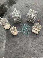 Overview of six assorted decorative bird cages on asphalt. Five are white metal cages with various designs; one is blue metal decorative wire object. All show rust and paint wear.