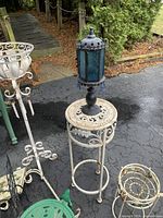Photo showing white metal tall round side table with a black and blue lantern on top, positioned on asphalt outdoor surface