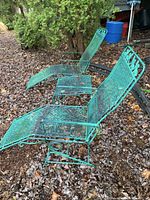 Two matching turquoise metal mesh lounge chairs with reclining and rocking features placed on leaf-covered ground outdoors near shrubbery and a house.