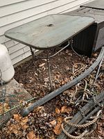 Side view of outdoor table showing metal frame and legs with rust and wear, surrounded by fallen leaves.