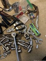 Overview of assorted hand tools including pliers, wrenches, sockets, ratchet, and small hardware parts on carpeted surface.
