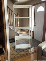White plastic shelving unit assembled with four shelves and four legs, positioned in a residential entryway near stairs and door.