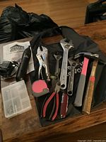 Photo showing assortment of hand tools including hammers, adjustable wrenches, locking pliers, measuring tape, JobMate cordless screwdriver and bits, and Swiss Army knife on a table.