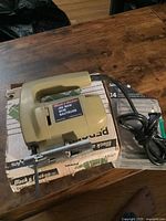 Black & Decker beige and black electric jigsaw tool on a cardboard box, cord visible, placed on wooden surface.