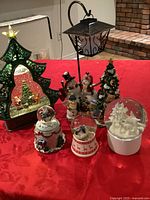 Photo of entire lot showing four snowglobes of varying sizes and a tree-shaped tea light ornament on a red tablecloth, illustrating scale and details of Christmas figures inside the globes and the ornament.