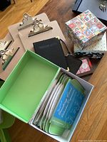 Eight wooden clipboards with metal clips, business card holder book, two decks of playing cards, pink coin purse, two nesting gift boxes, and greeting card organizer with assorted cards on a wooden floor.
