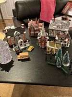 Full view of six lighted porcelain buildings arranged on table alongside small vehicles, trees, and benches.