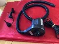 Electric air pump laying on red fabric, attached flexible hose and three nozzle adapters visible.