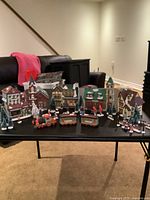 Full display of all porcelain Christmas village houses, train, trees and accessories on a black table in a living room setting.