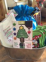 Holiday hostess gift lot packed in a metal tin displaying the tea towel, blue bag with snowball fight kit, green penguin potholders, and boxed Christmas tree bottle topper with charms visible.